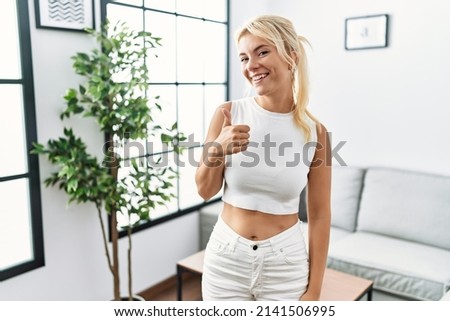 Young caucasian woman standing at living room doing happy thumbs up gesture with hand. approving expression looking at the camera showing success. 