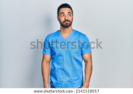 Handsome hispanic man with beard wearing blue male nurse uniform smiling looking to the side and staring away thinking. 