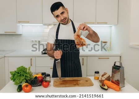 Portrait of a handsome man in the kitchen. A man dressed in a black apron. He looks at the camera. On the kitchen table are cooking products. Chicken. Vegetables. Spices