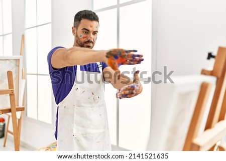 Young hispanic man looking canvas draw doing photo gesture with hands at art studio