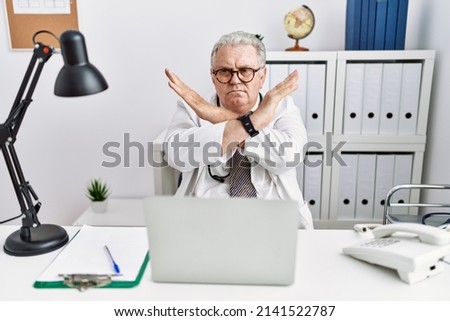 Senior caucasian man wearing doctor uniform and stethoscope at the clinic rejection expression crossing arms doing negative sign, angry face 