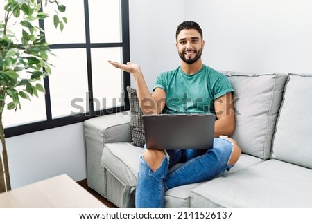 Young handsome man with beard using computer laptop sitting on the sofa at home smiling cheerful presenting and pointing with palm of hand looking at the camera. 