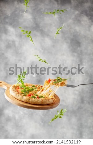 Levitation. Pizza with melted cheese, mozzarella, tomatoes, arugula and jalapeno pepper. Italian pizza on a wooden cutting board. Flying food. A slice of pizza on a shovel with stretching cheese. Grey Royalty-Free Stock Photo #2141532119