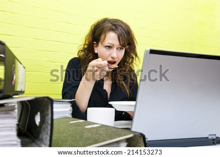 Caucasian young woman eating and working on her laptop computer at her desk