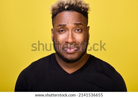 An upset guy in a black T-shirt crooked his face, looking into the camera with sad eyes. Against a yellow background, a sad dark-skinned man with curly hair almost burst into tears with resentment. Royalty-Free Stock Photo #2141536655