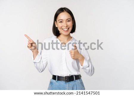 Image of young asian business woman, smiling while pointing finger left and showing thumbs up, recommending product, praise, standing over white background