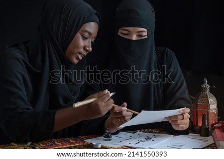 Muslim women wearing black hijab learn to writing Arabic calligraphy with ink together, smiling and looking each other, black background, Arabic letters mean the name of Muslim god "Allah"