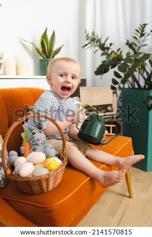 A little smiling boy sitting on the orange sofa  with easter bunny, watering can and basket with eggs - preschool kid