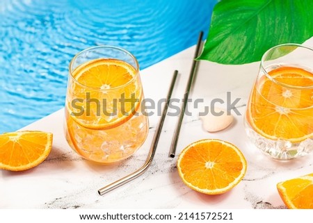 Summer refreshing water with orange slice. Glass of cocktail, orange slice, metal straw and tropical monstera leaf on beach swimming pool. Summer relaxation background.
