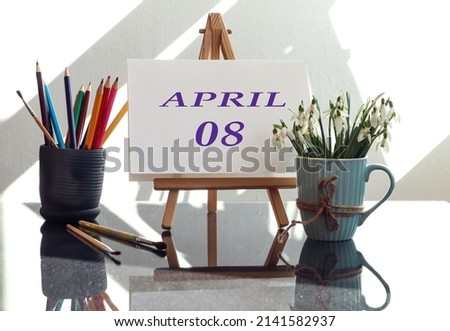 Calendar for the eighth of April: an easel with the inscription April in English and the numbers 08, a bouquet of snowdrops in a cup, multi-colored pencils, brushes in the sun from the window