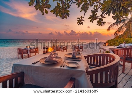 Outdoor restaurant at the beach. Table setting at tropical beach restaurant. Led light candles and wooden tables, chairs under beautiful sunset sky, sea view. Luxury hotel or resort restaurant Royalty-Free Stock Photo #2141601689