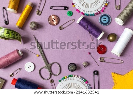 Frame with sewing tools. Sewing threads, needles, pins, scissors, buttons on a purple background with a place for text. Top view, Royalty-Free Stock Photo #2141632541