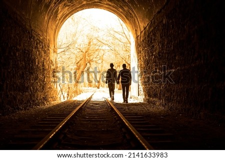 Couple walking hand in hand along the track through a railway tunnel towards the bright light at the other end, they appear as silhouettes against the light. back view. Royalty-Free Stock Photo #2141633983