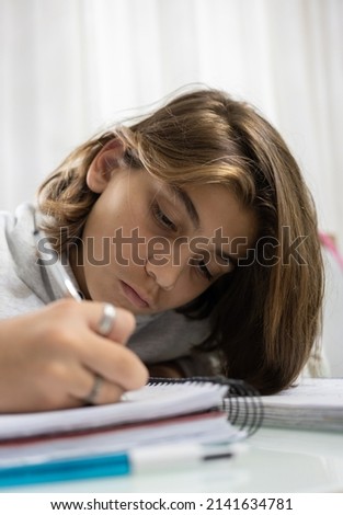 
Boy in the background, with white skin and long hair, writing in his notebook. Studying and doing school homework.