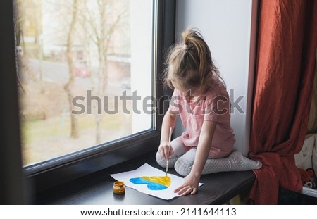 A little sad Ukrainian girl, being far from home, draws the flag of Ukraine in the form of a heart on the windowsill. War and children.