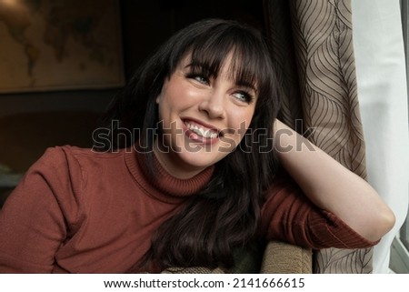 Portrait of a caucasian woman in her 30s smiling while sitting on the couch near a window.  Royalty-Free Stock Photo #2141666615
