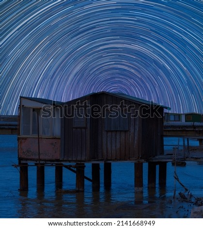 Star trails in the night sky in Italy. Movement of the stars behind an old metal and wooden fishing shed standing in the water