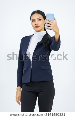 Studio shot Asian young beautiful female professional successful businesswoman model in formal business suit and turtleneck shirt standing holding smartphone taking selfie photo on white background.