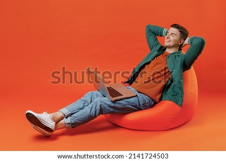 Full size body length young brunet man 20s wears red t-shirt green jacket sit in bag chair hold use work on laptop pc computer relax eyes closed isolated on plain orange background studio portrait.