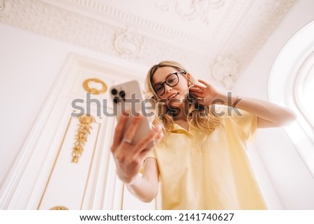 Young caucasian cheerful woman using mobile phone in bright living room, connecting to friends in video call or taking selfie photo.