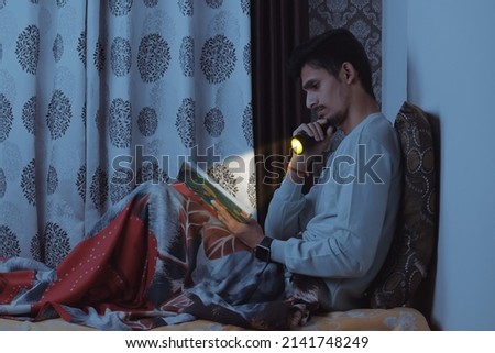 Young bearded Indian boy sitting on bed in his bedroom and reading book very seriously at night with tourch wearing light blue tshirt and red bedsheet isolated on textured curtain background
         