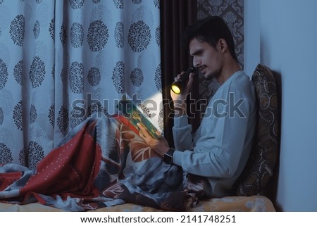 Young bearded Indian boy sitting on bed in his bedroom and reading book very seriously at night with tourch wearing light blue tshirt and red bedsheet isolated on textured curtain background
         