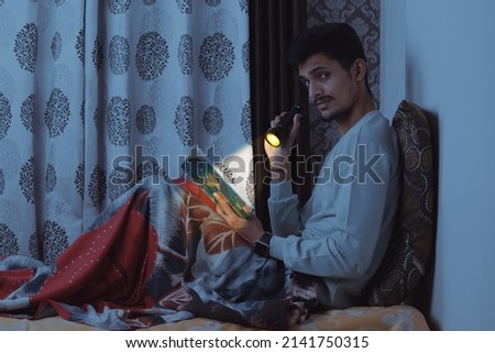 Young bearded Indian boy sitting on bed in his bedroom and reading very scary and horror book at night with tourch wearing light blue tshirt and red bedsheet isolated on textured curtain background
