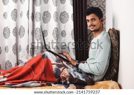 Young smiling bearded Indian boy sit on bed in bedroom makes video call looks at camera conversation by distant video call, distance hiring job interview process tutor and trainee study online concept Royalty-Free Stock Photo #2141759237