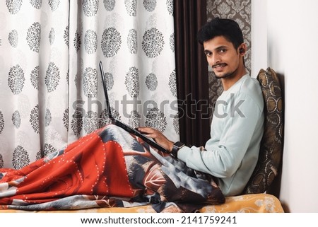 Young smiling bearded Indian boy sit on bed in bedroom makes video call looks at camera conversation by distant video call, distance hiring job interview process tutor and trainee study online concept Royalty-Free Stock Photo #2141759241