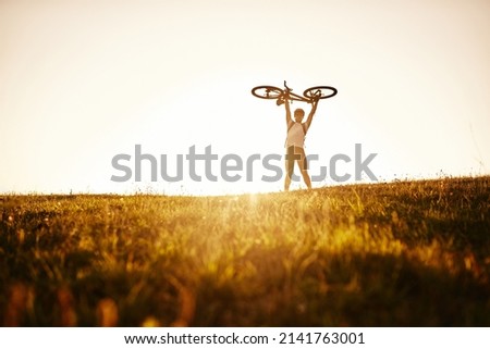 The cyclist is holding his bicycle over himself on the background of red sunset. Biker with bicycle on the field during sunrise. High quality photo Royalty-Free Stock Photo #2141763001