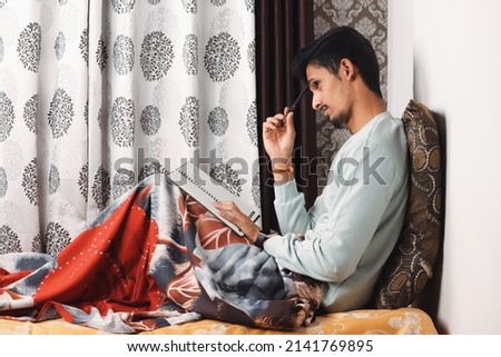 Young bearded Indian Boy thinking about what to write in note book in light blue t-shirt seating and deep thinking expression isolated on textured curtain studio background concept story writing
