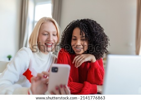 Happy smiling young women friends looking at photos on mobile phone. Girls havin fun. Two Female Friends Relaxing At Home Talking Together