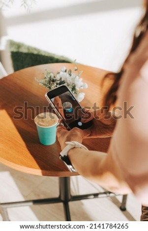 A woman takes pictures of a delicious cappuccino on a smartphone. Aromatic coffee to go. Hands remove coffee for social networks