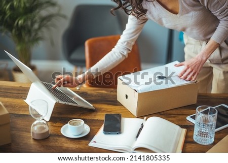 A Caucasian business woman smiles and uses a laptop to check the information on the package shipping box before sending it to the customer. A small entrepreneur who works at home.