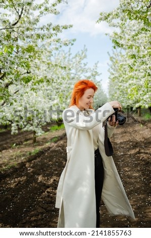 Girl photographer with red hair with a camera. female tourist, blogger, photographs nature. spring background. concept of work, hobby, tourism. spring light green tones. Royalty-Free Stock Photo #2141855763