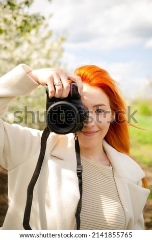 Girl photographer with red hair with a camera. female tourist, blogger, photographs nature. spring background. concept of work, hobby, tourism. spring light green tones. Royalty-Free Stock Photo #2141855765