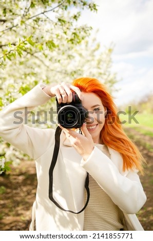Girl photographer with red hair with a camera. female tourist, blogger, photographs nature. spring background. concept of work, hobby, tourism. spring light green tones. Royalty-Free Stock Photo #2141855771