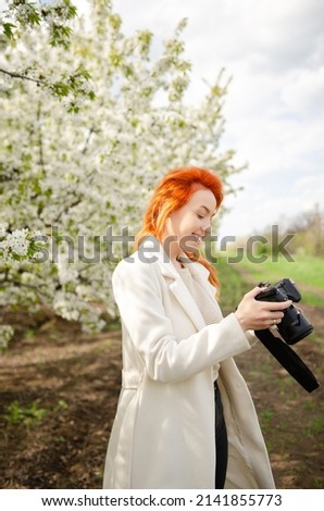 Girl photographer with red hair with a camera. female tourist, blogger, photographs nature. spring background. concept of work, hobby, tourism. spring light green tones. Royalty-Free Stock Photo #2141855773