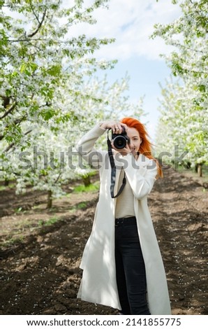 Girl photographer with red hair with a camera. female tourist, blogger, photographs nature. spring background. concept of work, hobby, tourism. spring light green tones. Royalty-Free Stock Photo #2141855775