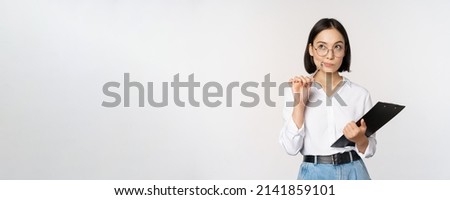 Asian girl in glasses thinks, holds pen and clipboard, writing down, making notes, standing over white background Royalty-Free Stock Photo #2141859101