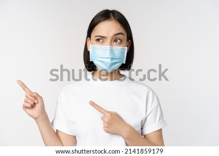 Image of asian woman in medical mask from covid, looking confused left, pointing at logo, showing advertisement, standing over white background