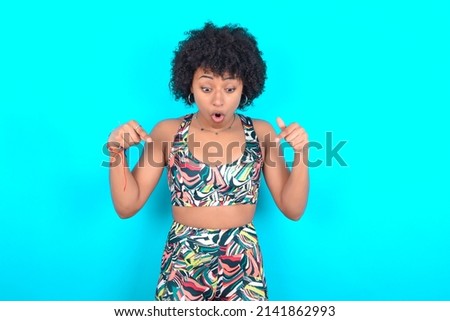 Amazed young woman with afro hairstyle in sportswear against blue background points down with fore fingers, opens mouth being shocked. Advertisement concept.