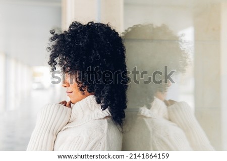 Side view of a relaxed young woman with curly hair in white sweater leaning on glass touching her chin looking ahead.