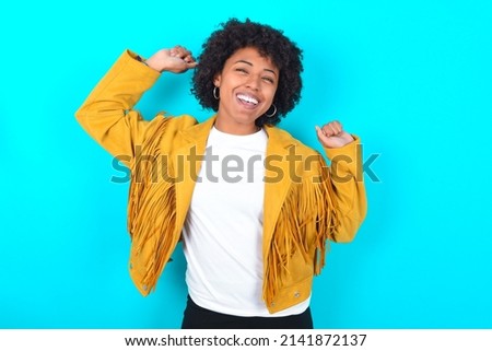 Photo of upbeat Young woman with afro hairstyle wearing yellow fringe jacket over blue background has fun and dances carefree wear being in perfect mood makes movements. Spends free time on disco 