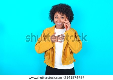 Smiling Young woman with afro hairstyle wearing yellow fringe jacket over blue background talks via cellphone, enjoys pleasant great conversation. People, technology, communication concept