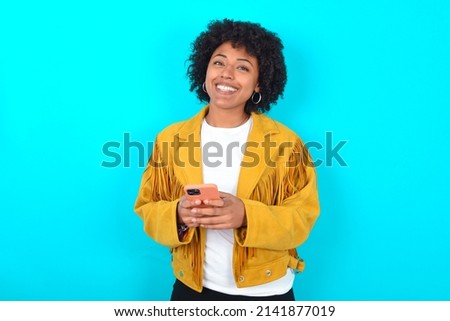 Young woman with afro hairstyle wearing yellow fringe jacket over blue background Mock up copy space. Using mobile phone, typing sms message