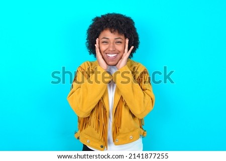 Happy Young woman with afro hairstyle wearing yellow fringe jacket over blue background touches both cheeks gently, has tender smile, shows white teeth, gazes positively straightly at camera,