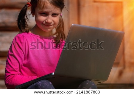 Learn always and everywhere. Portrait of young beautiful school girl uses a laptop and studies remotely outdoorson the background of a wooden house. Horizontal image.