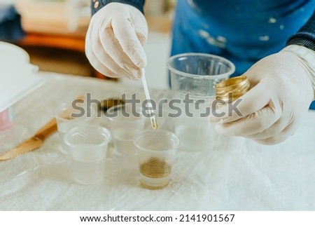 Artist prepairing ingredients for create clock in Resin art technique. Woman wearing rubber gloves mixing golden mica powdered pigment and medium. Selective focus. Royalty-Free Stock Photo #2141901567