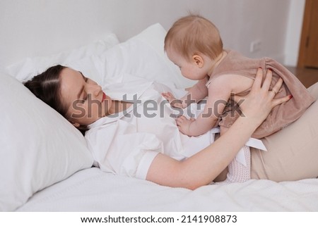 Sweet young mother playing with her little 1-year-old daughter in bed.
Caring brunette woman laying with her baby child at home. Tenderness and love between mother and daughter. Lovely family indoors.